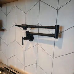 Matte black wall-mount pot filler faucet with swivel brass arm on tiled backsplash.