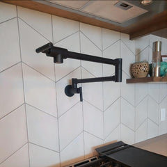 Matte black wall-mount pot filler faucet with swivel brass arm on white tiled backsplash.