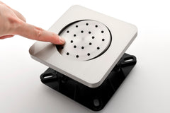 Silver square shower valve with circular perforated center, black base, and hand touching it.