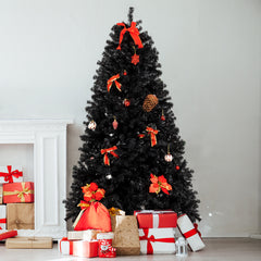A 7-foot black PVC Christmas tree with 1500 dense branches, decorated with red ribbons, ornaments, pinecones, and poinsettias, standing on an iron base beside a fireplace and wrapped gifts.