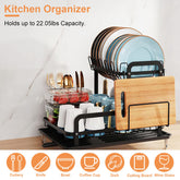 2-tier black carbon steel dish drying rack with drainboard, holding dishes, utensils, cups, and a cutting board.