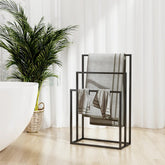 Black iron 3-tier freestanding towel rack with towels, beside a bathtub and plant.