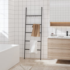 Black 5-tier iron towel ladder rack leaning in a white tiled bathroom with towels draped on rungs.