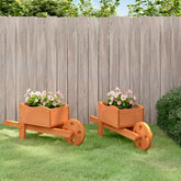 Two wooden wheelbarrow planters with pink flowers, placed on grass against a fence.