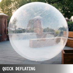 Large transparent inflatable zorb ball deflating on a paved outdoor area near greenery.