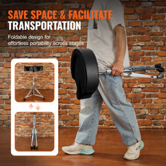 A man carries a folded adjustable saddle drum throne with black seat and tripod legs against a brick wall.