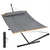 A gray quilted two-person hammock with a pillow, wooden spreader bars, and a black steel stand.