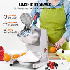 Silver stainless steel electric ice crusher with ice cubes and shaved ice on a table.