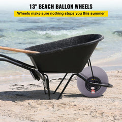 Two 13-inch black TPU balloon wheels with gray hubs for a kayak dolly, shown attached to a black wheelbarrow on a sandy beach.