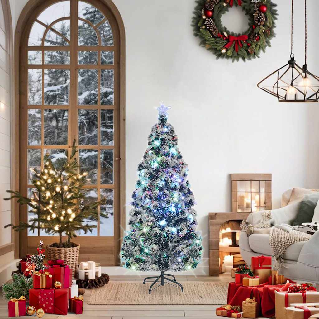 A 5 ft pre-lit fiber optic Christmas tree in green and white, twinkling with colorful lights, displayed in a cozy holiday living room.