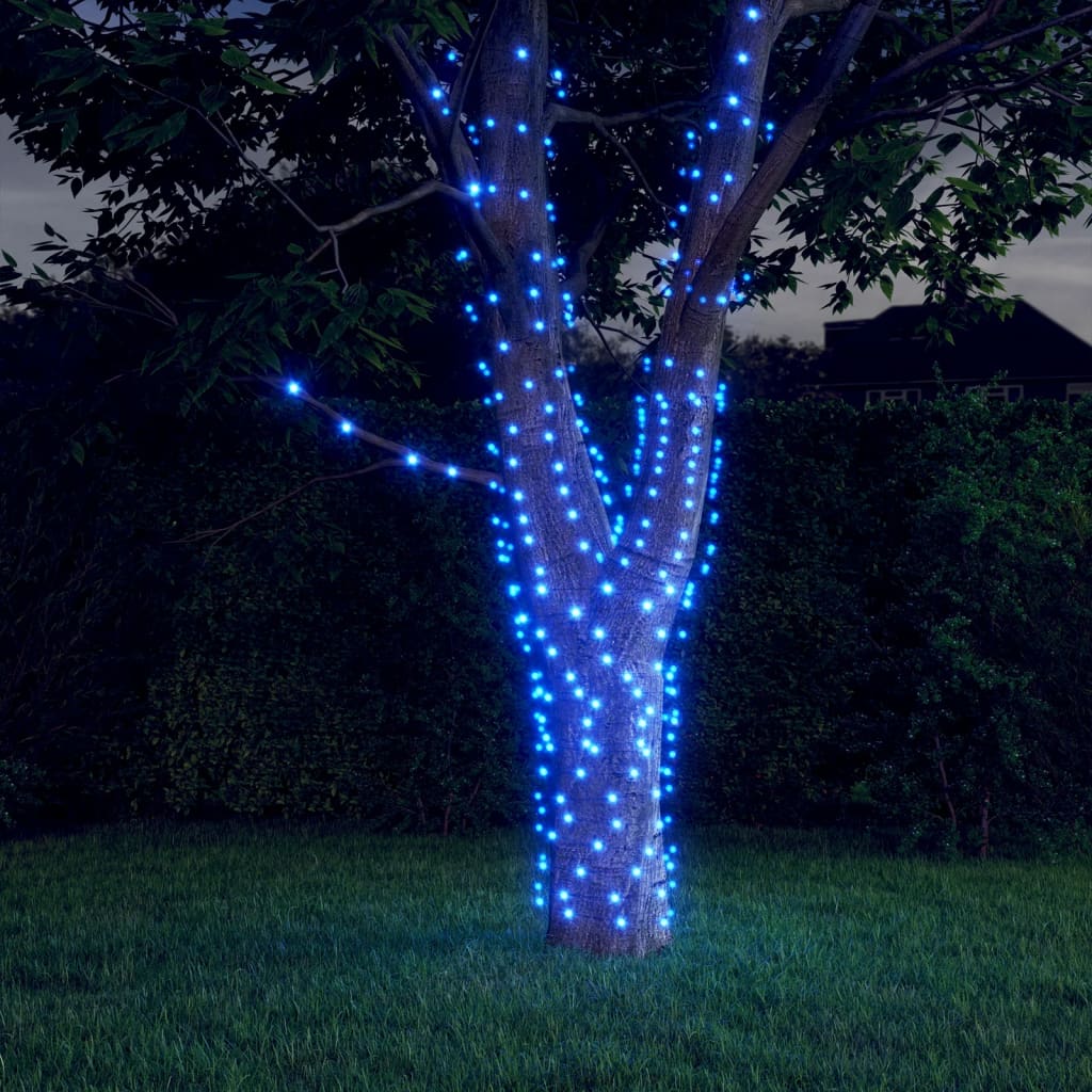 A tree wrapped in two strands of blue solar fairy lights glowing at dusk in a yard.